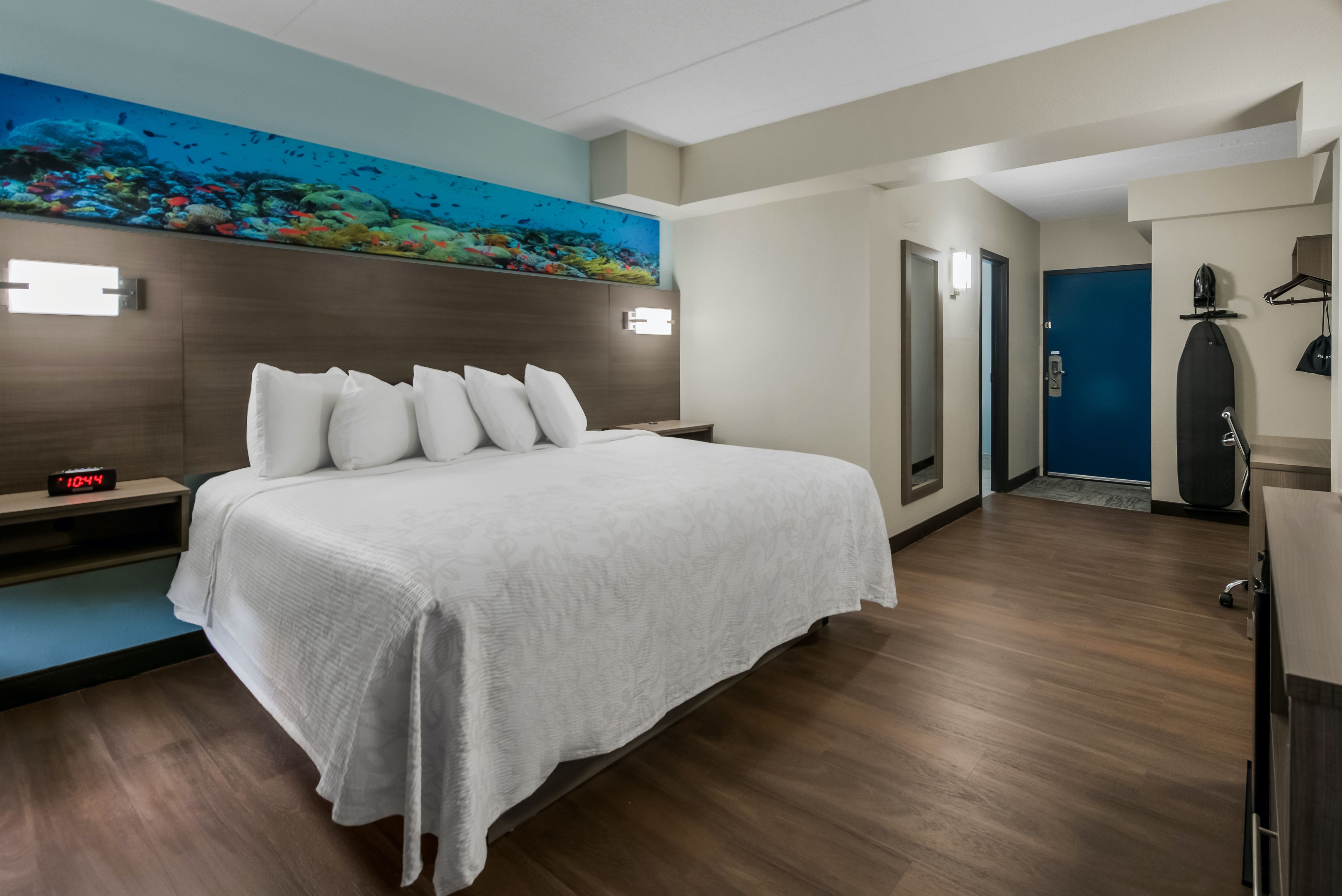 Quarto de hotel com cama, mesa de cabeceira, relógio de parede, pintura de uma cena subaquática e porta para outro cômodo.