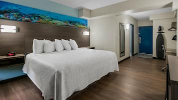 Quarto de hotel com cama, mesa de cabeceira, relógio de parede, pintura de uma cena subaquática e porta para outro cômodo.