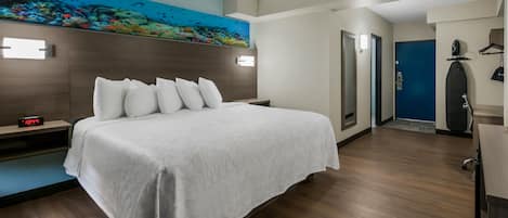 A hotel room with a bed, a nightstand, a wall clock, a painting of an underwater scene, and a door leading to another room.