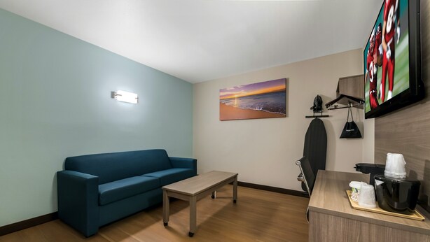 Quarto de hotel com sofá azul, uma mesa pequena, uma pintura de praia, escrivaninha com cafeteira e televisão na parede.