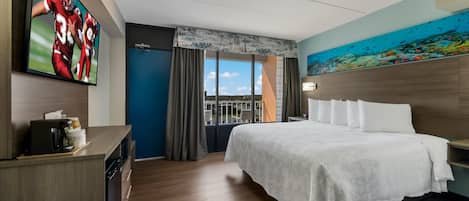 A hotel room with a large bed, a television on the wall, and a painting of an underwater scene.