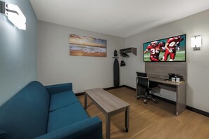Quarto com sofá azul, mesa de madeira, escrivaninha com computador, televisão e pintura de uma praia.