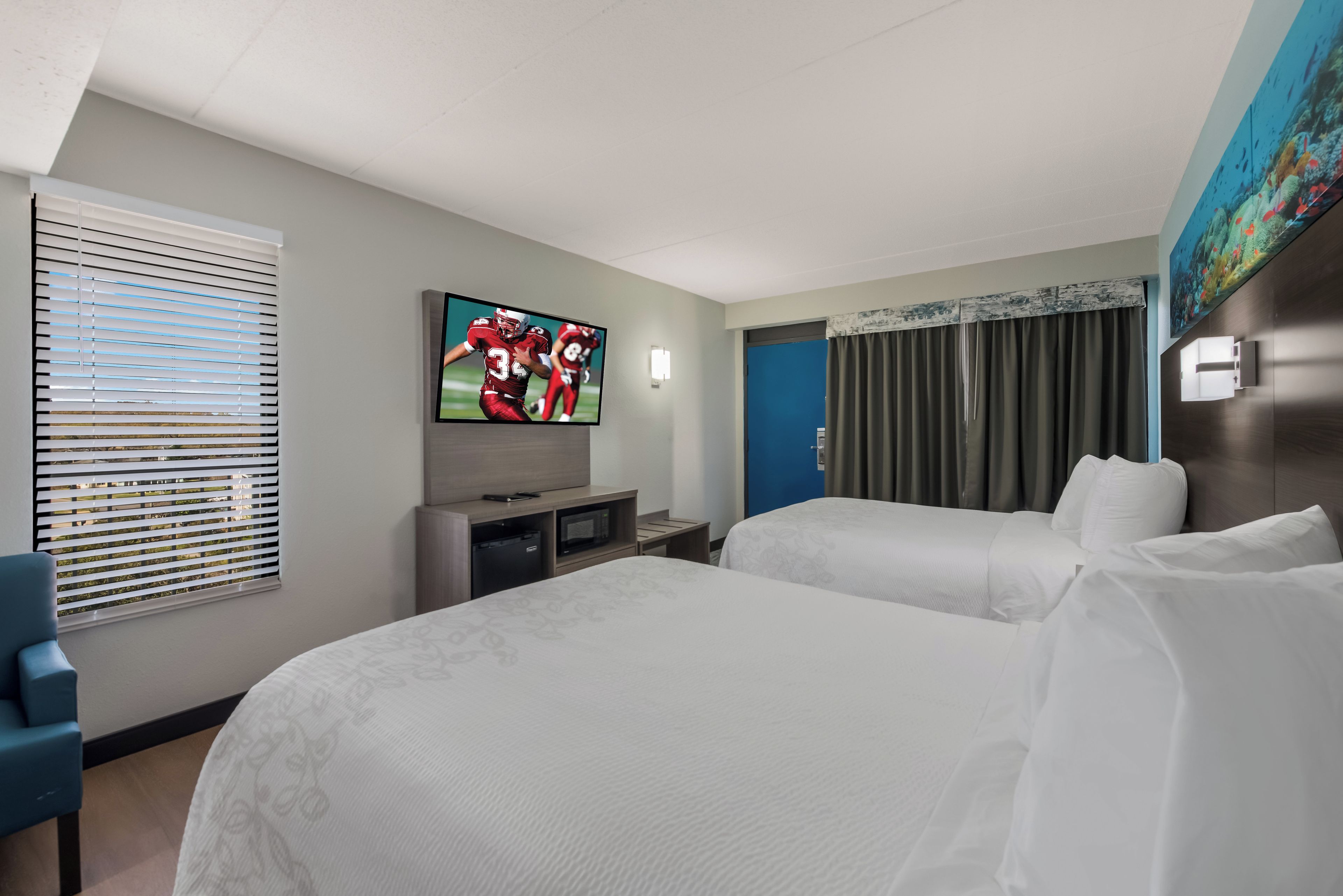A hotel room with two beds, a television, a desk, and a window with blinds.