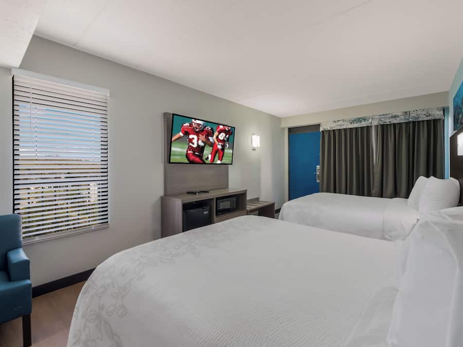 A hotel room with two beds, a television, a desk, and a window with blinds.