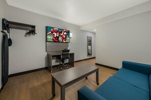 A modern living room with a flat-screen TV, a desk, a chair, a coffee table, and a blue sofa. - Red Roof Inn PLUS+ & Suites Virginia Beach – Seaside (Virginia Beach)