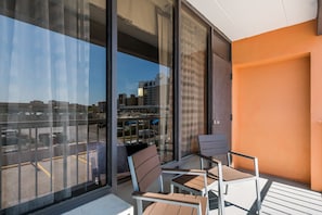 A balcony with two chairs and a table, a glass door, and a view of a parking lot and buildings. - Red Roof Inn PLUS+ & Suites Virginia Beach – Seaside (Virginia Beach)