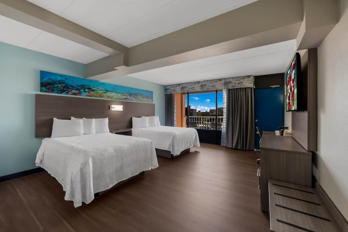 Habitación de hotel con dos camas, un ventanal con vista a la ciudad, un cuadro en la pared y un televisor de pantalla plana.