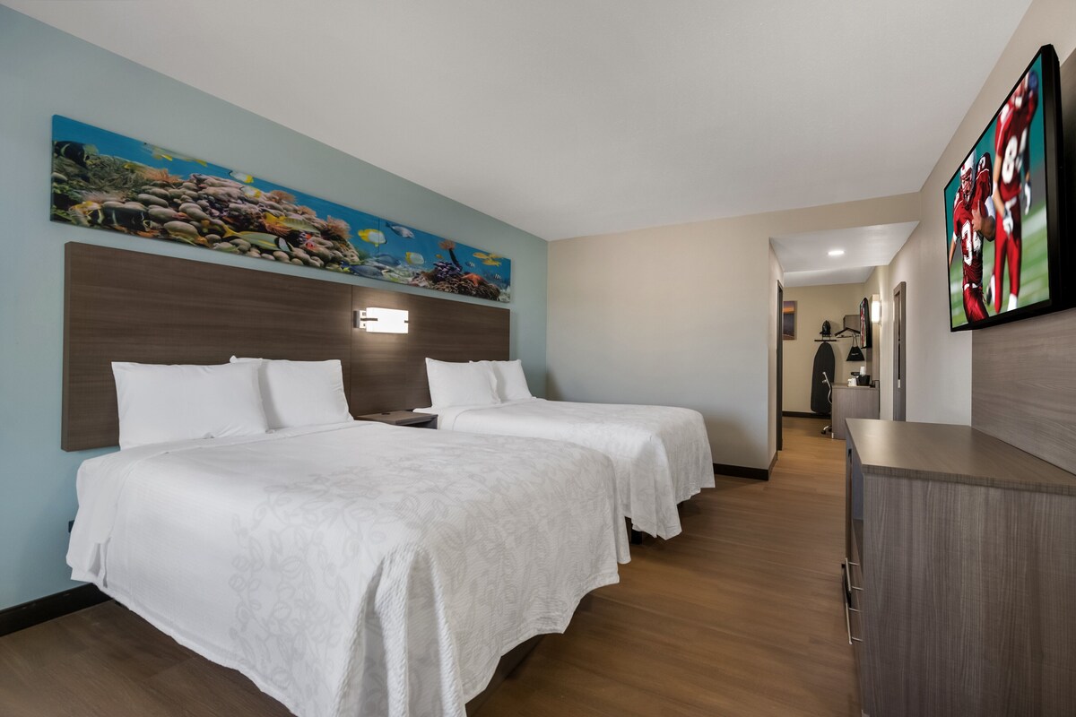 A hotel room with two beds, a large painting of an underwater scene, a flat-screen TV, and a wooden dresser.