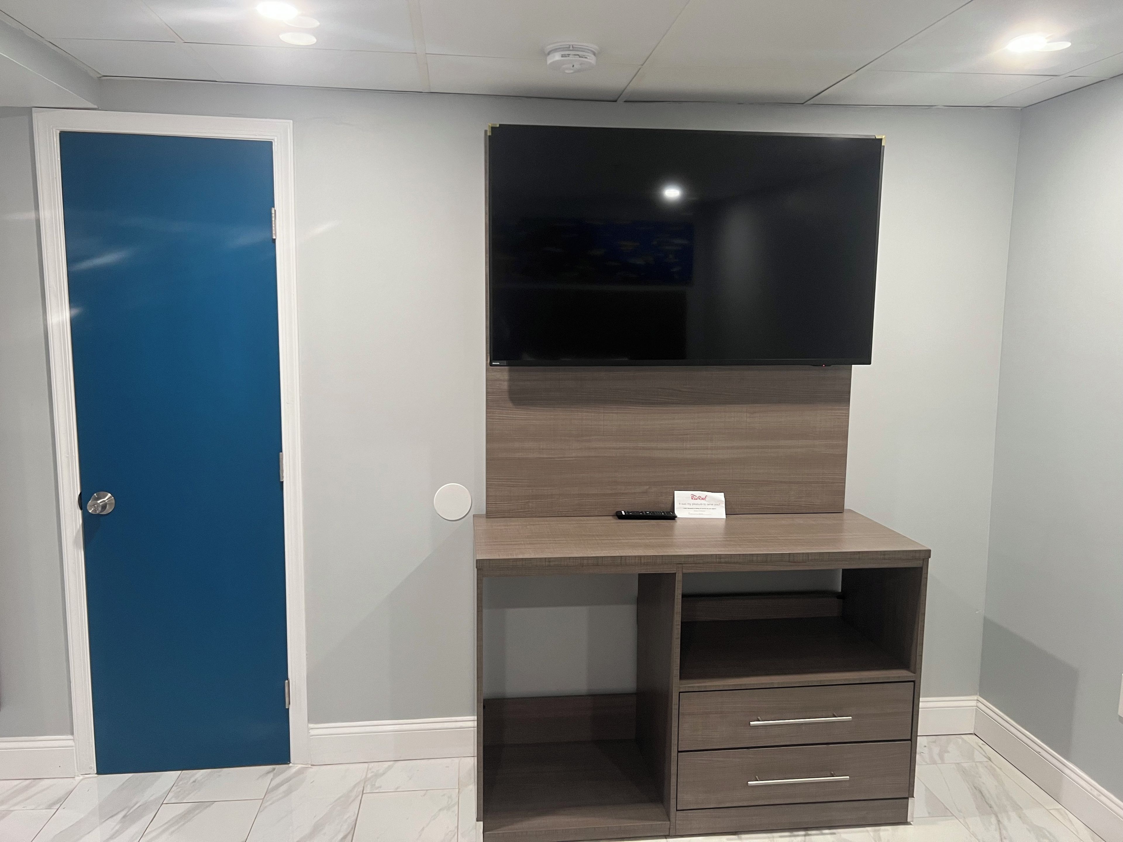 A room with a blue door, a wooden desk with drawers, a mounted flat-screen TV, and a white ceiling with recessed lighting.