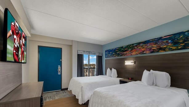 A hotel room with two beds, a TV, a painting, and a view of the city. - Red Roof Inn PLUS+ & Suites Virginia Beach – Seaside (Virginia Beach)
