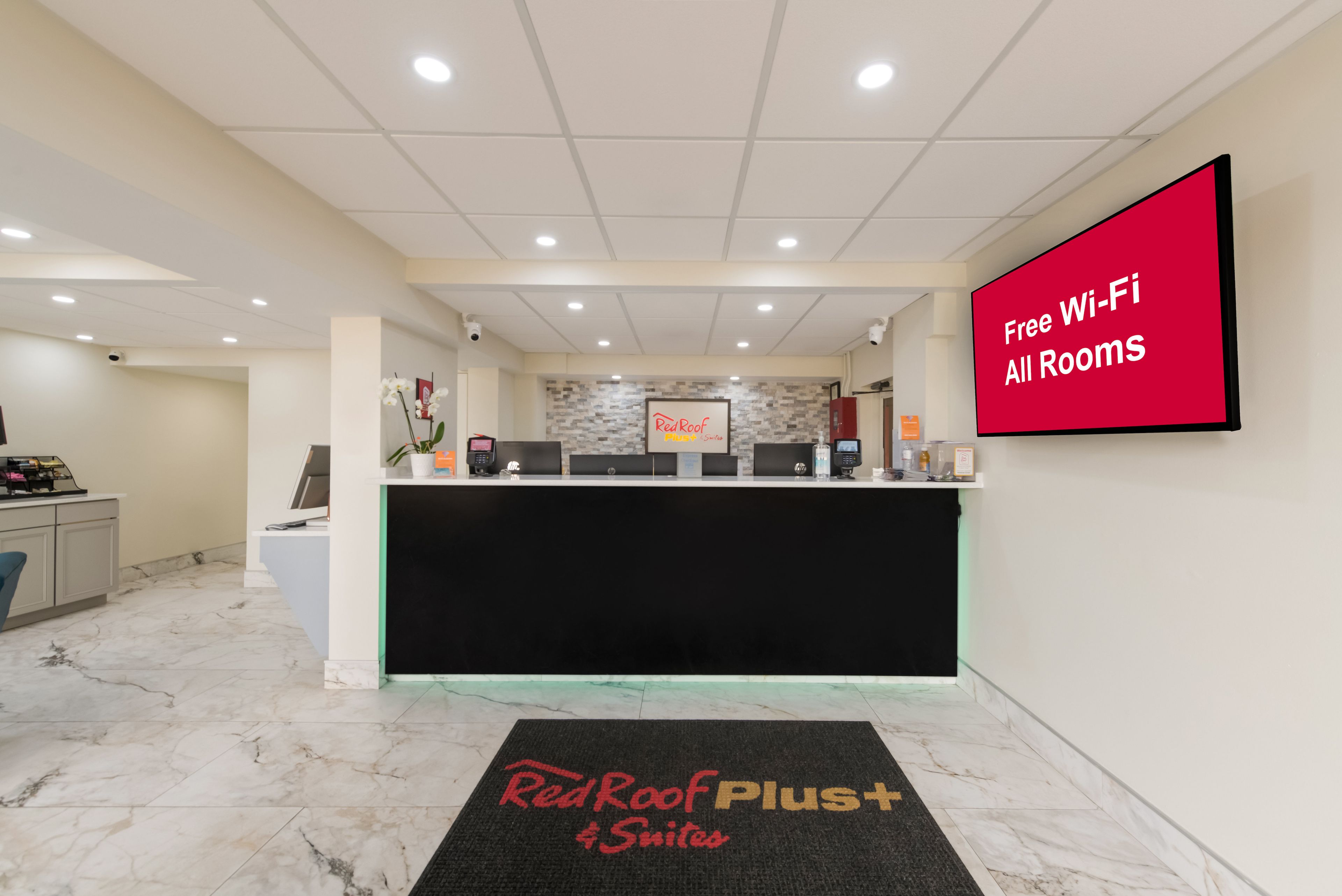 A hotel reception area with a Red Roof Plus+ sign, a Red Roof Plus+ sign on the wall, and a Red Roof Plus+ sign on the floor mat.