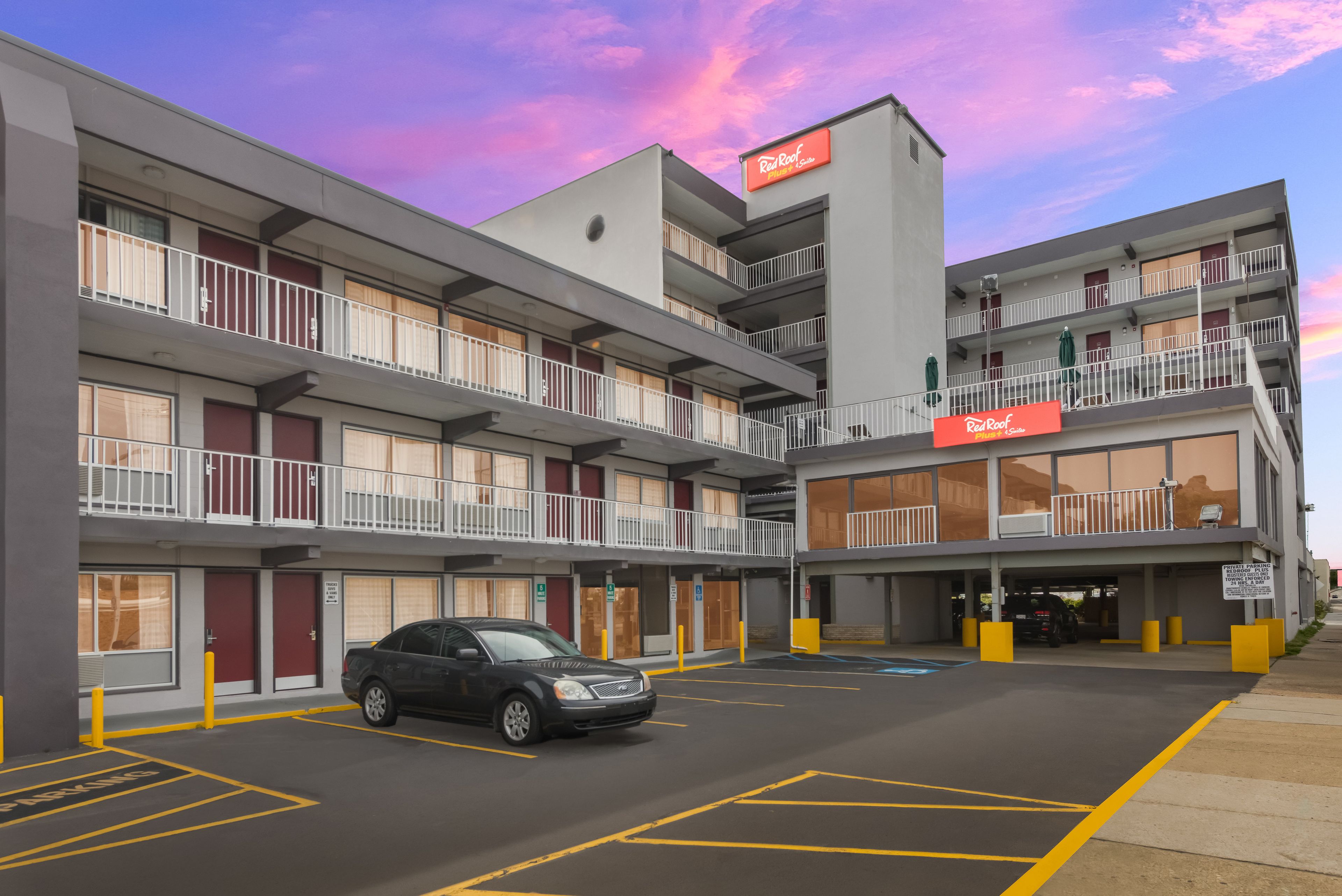 A multi-story hotel with a parking lot, a sign for Red Roof Plus & Suites, and a black car parked in the lot.