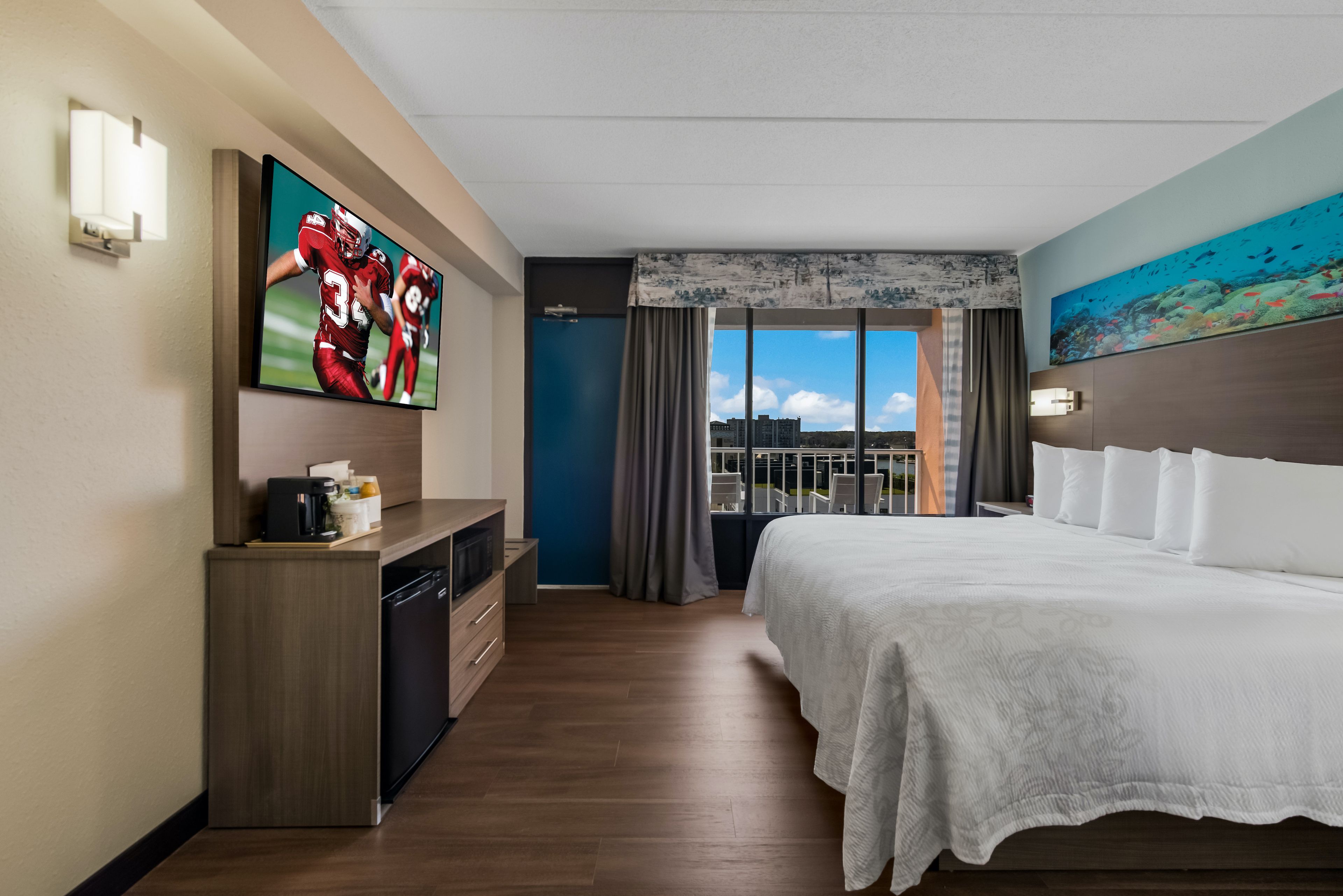 Ein Hotelzimmer mit einem großen Bett, einem an der Wand befestigten Fernseher, einem Schreibtisch mit Kaffeemaschine und Blick auf die Stadt durch das Fenster.