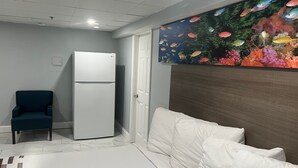 Quarto com cama, parede branca, porta, geladeira branca e cadeira azul.