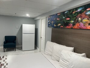A bedroom with a bed, a white wall, a door, a white refrigerator, and a blue chair. - Red Roof Inn PLUS+ & Suites Virginia Beach – Seaside (Virginia Beach)