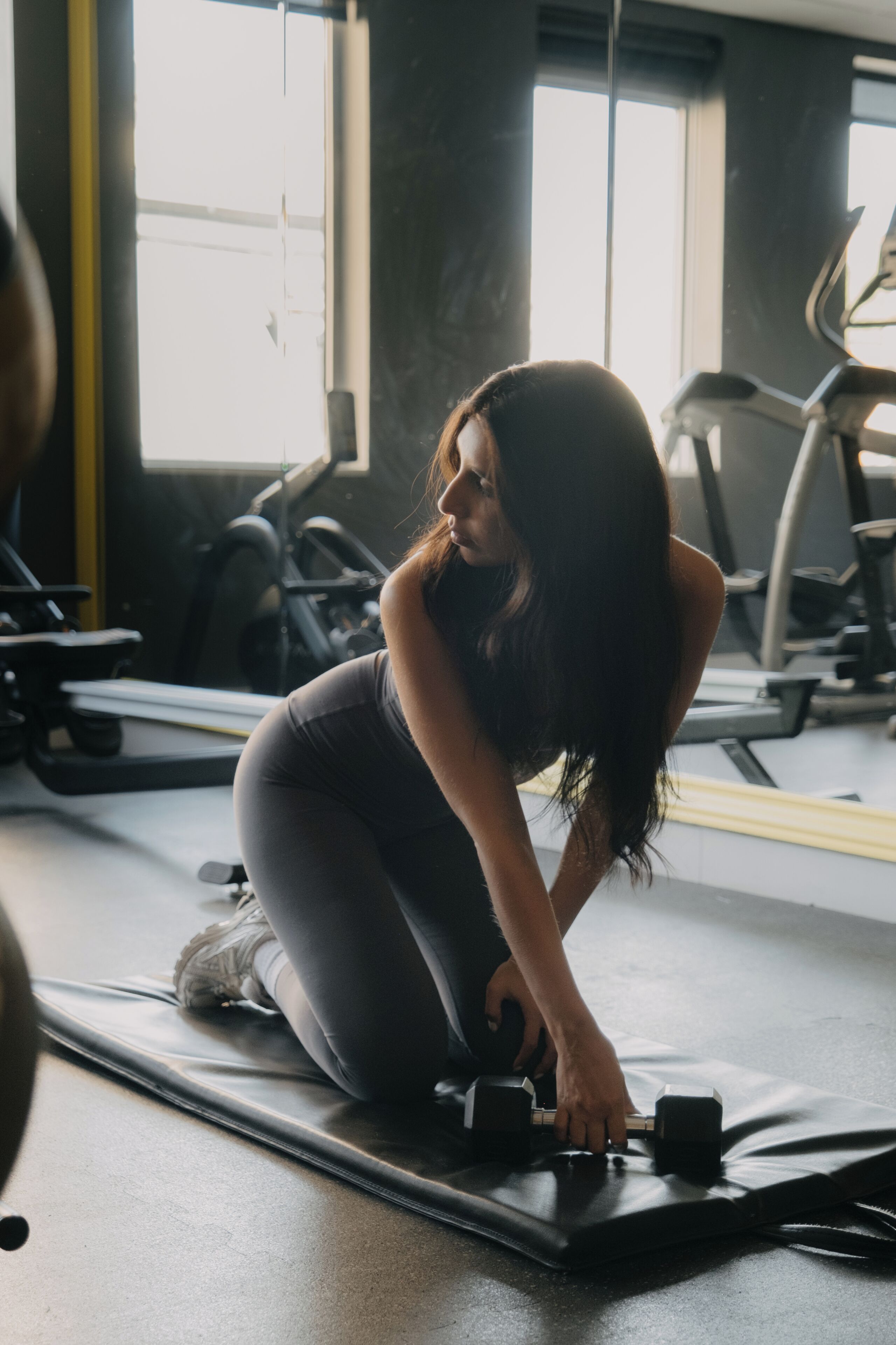 Una persona ejercitándose sobre una colchoneta con mancuernas, en un gimnasio con equipo de ejercicio al fondo.