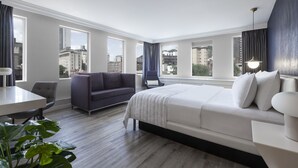 A modern hotel room with a large bed, a sofa, a desk, and a view of the city. - SENS Hotel (Montreal)