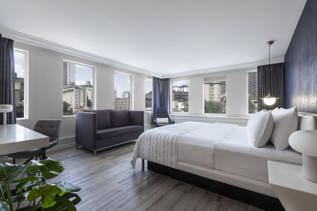 A modern hotel room with a large bed, a sofa, a desk, and a view of the city.