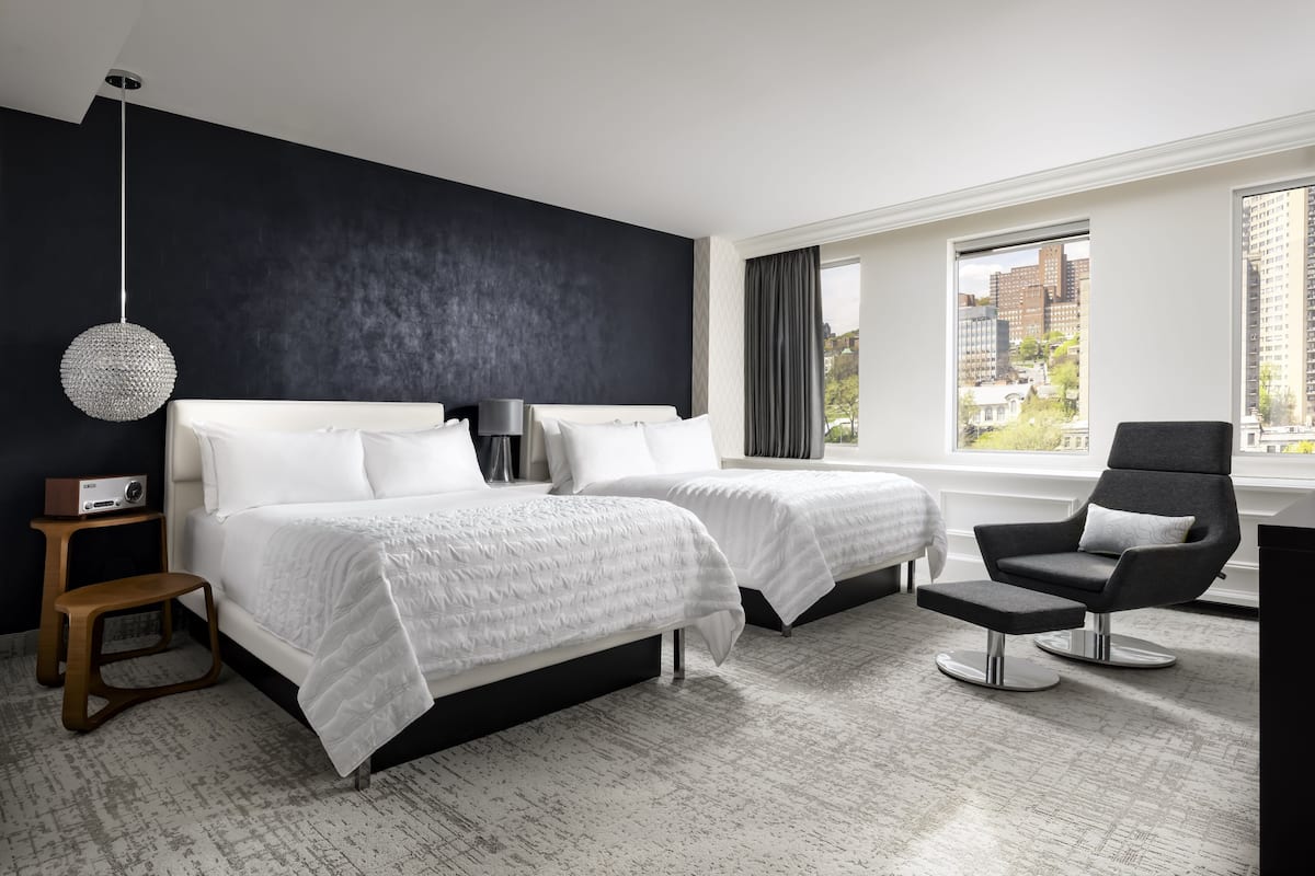 A modern hotel room with two beds, a black chair, a small table, and a city view.