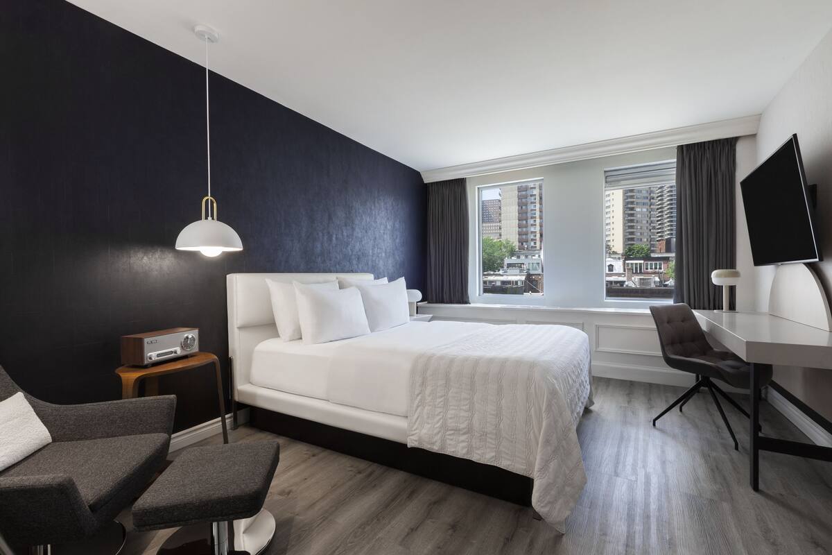 A modern hotel room with a large bed, a desk, a chair, a TV, and a window with a city view.