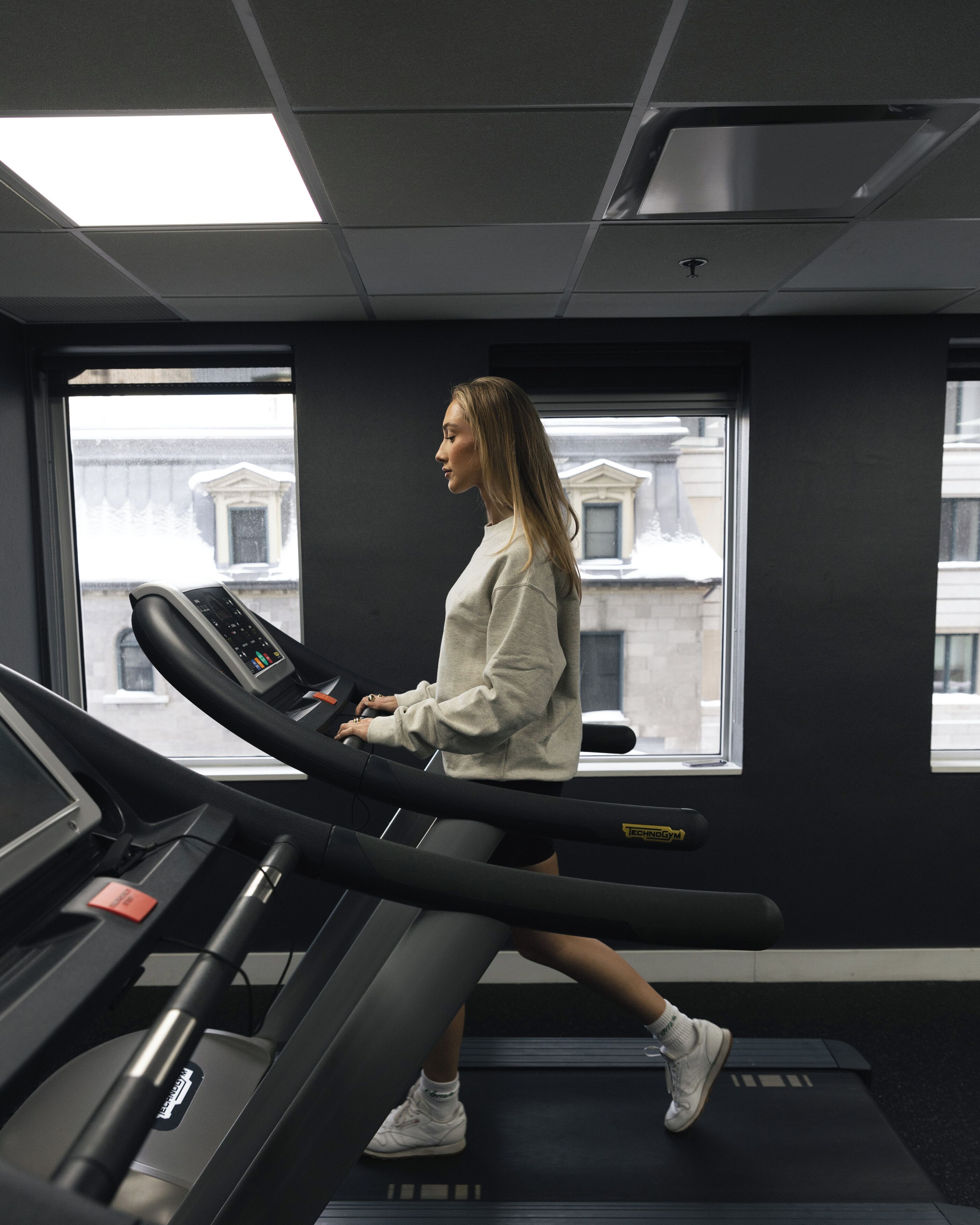 A person using a treadmill in a room with large windows, a visible cityscape, and a ceiling with recessed lighting.
