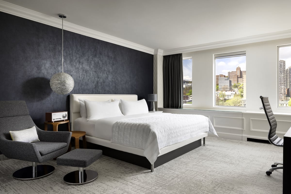 Quarto de hotel moderno com uma cama grande, uma poltrona cinza, uma pequena mesa, uma escrivaninha e vista para a cidade.