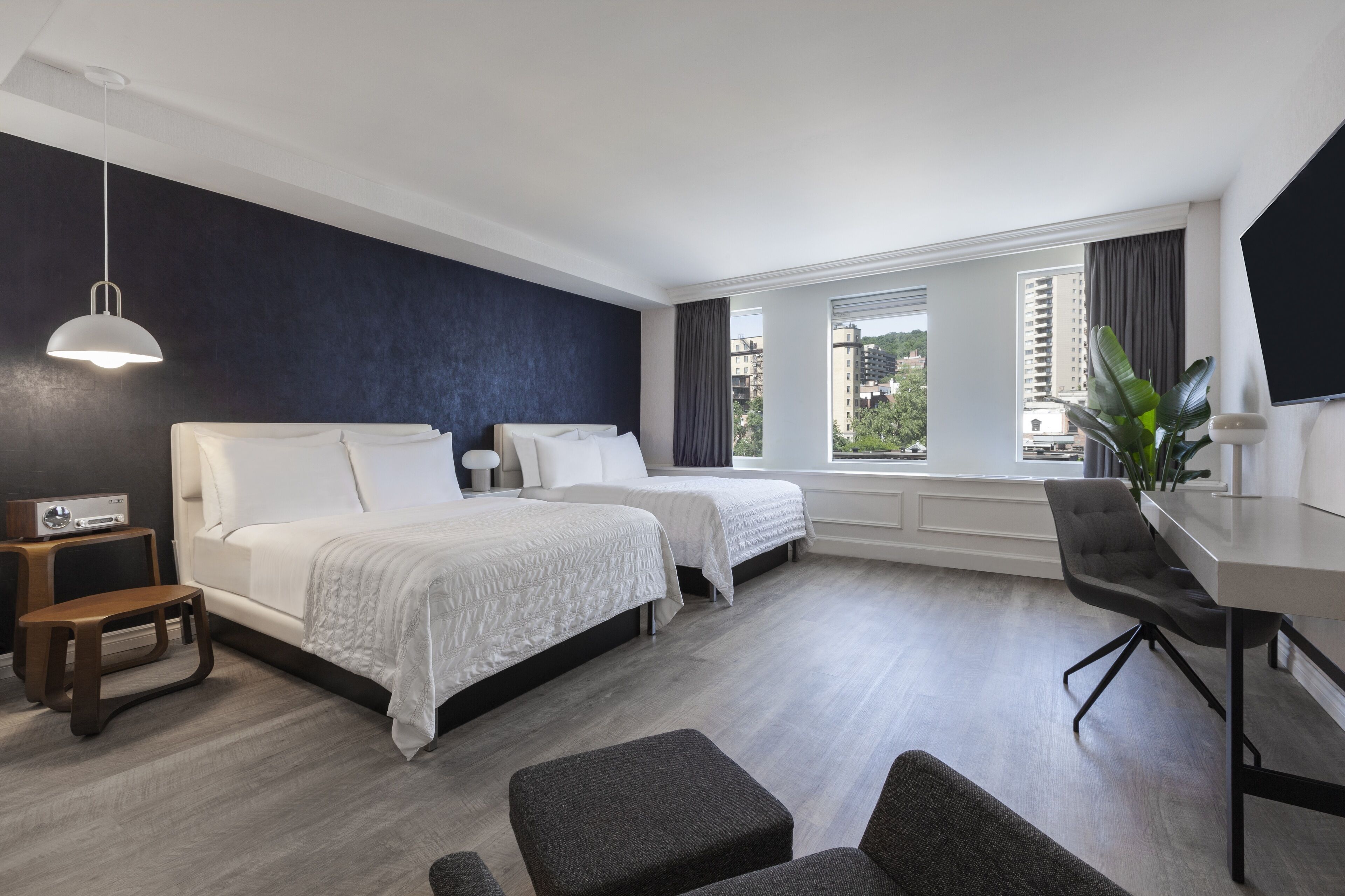 A modern hotel room with two beds, a desk, and a large window with a city view.