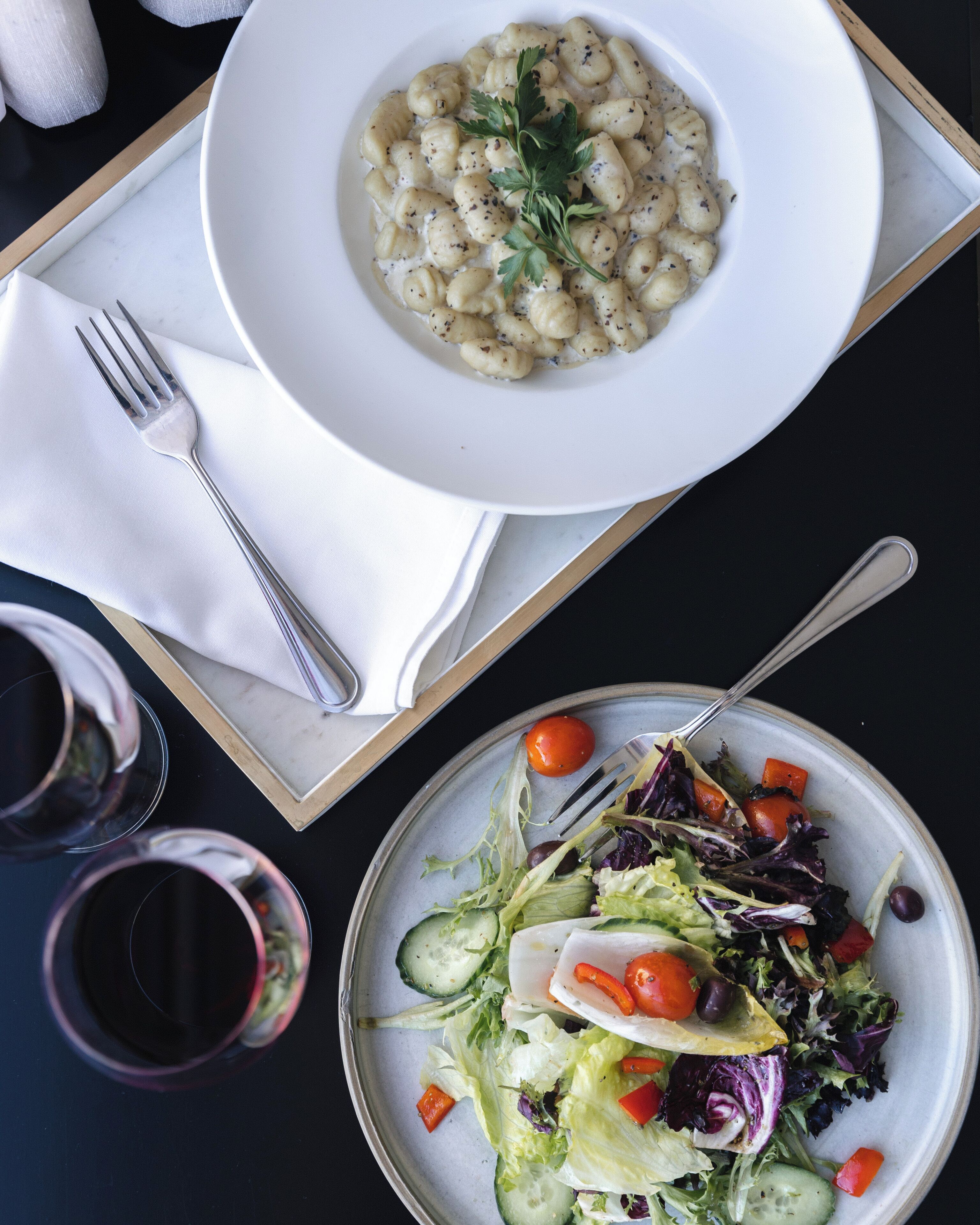 A dining table with a plate of gnocchi and a salad, a fork, and a glass of red wine.