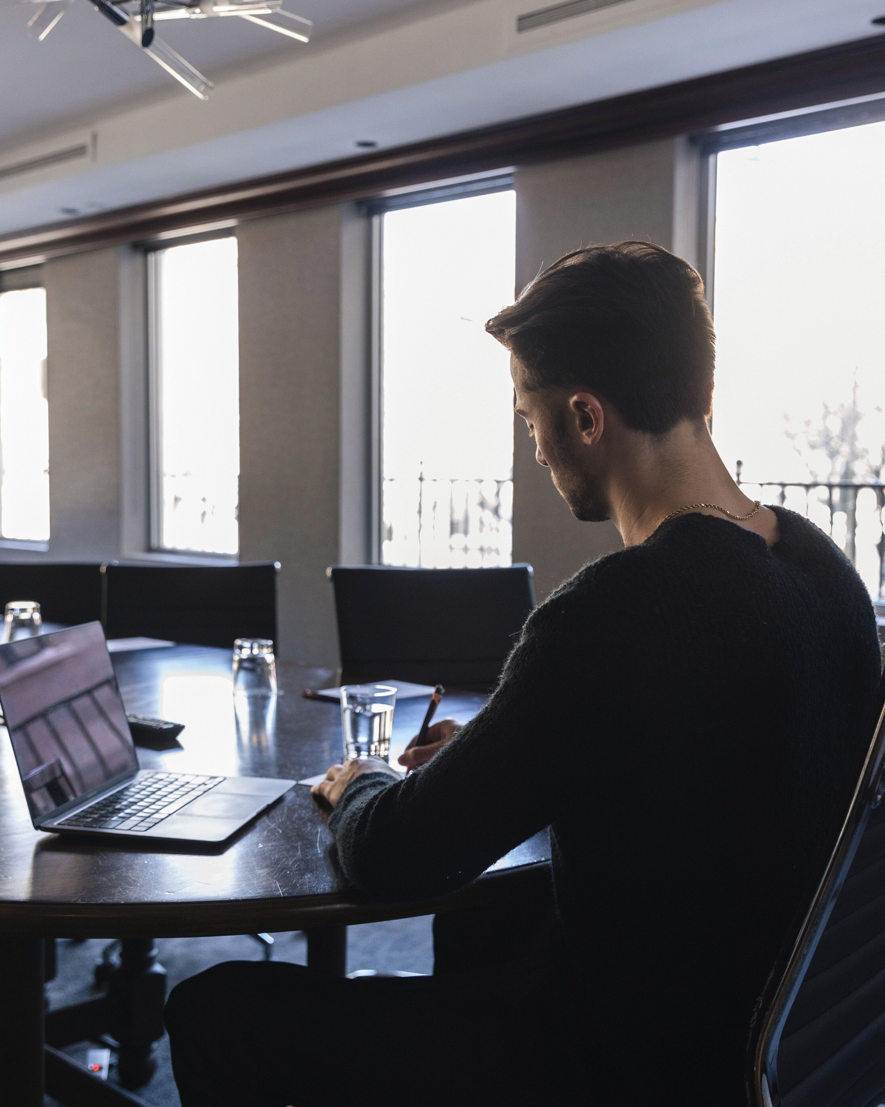 A person seated at a table with a laptop, writing, and a glass of water.