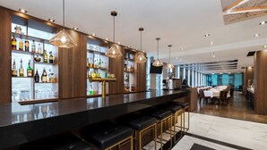 A modern bar area with a long counter, multiple stools, and shelves stocked with liquor bottles. - SENS Hotel (Montreal)