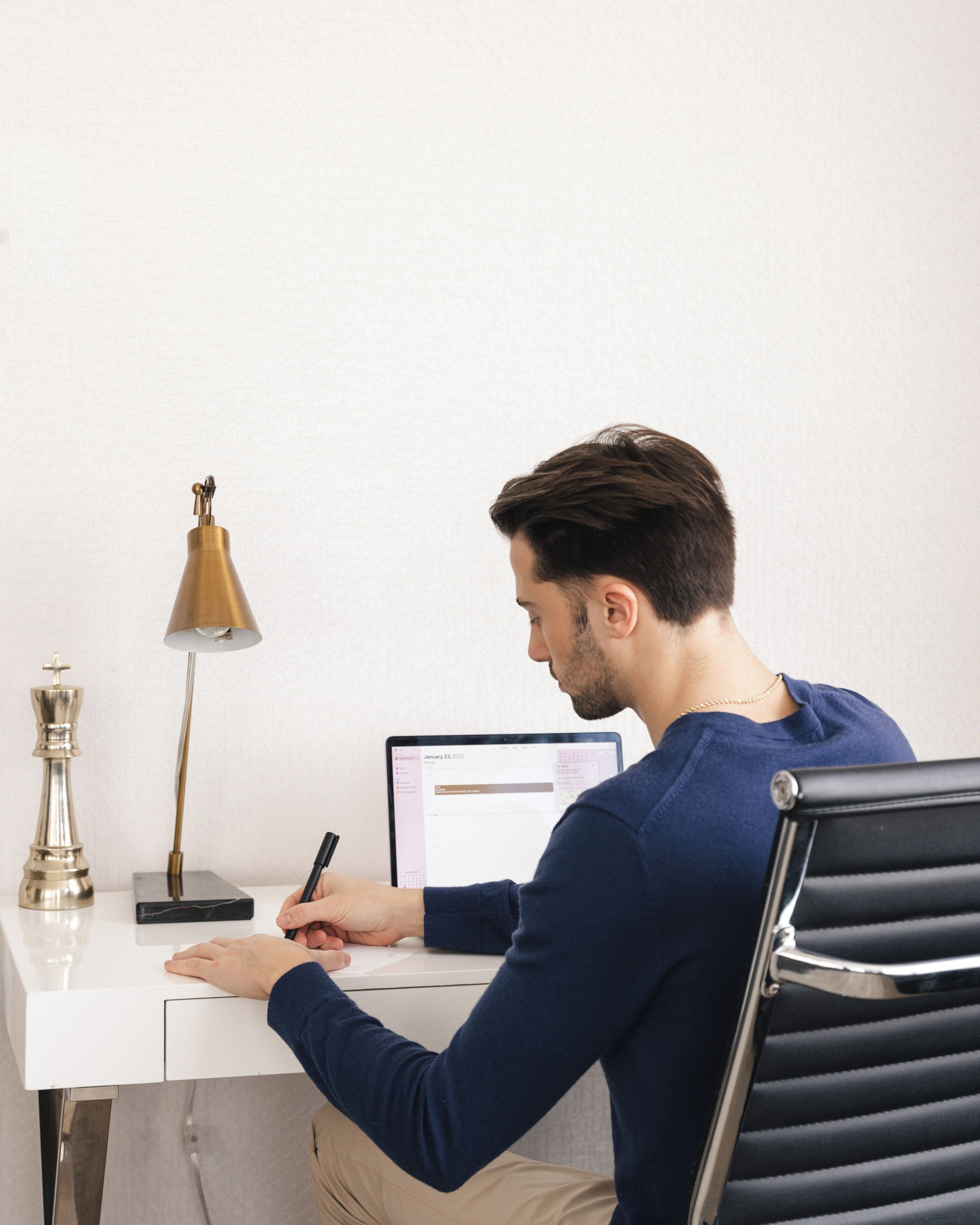 Eine Person sitzt an einem Schreibtisch mit einem Laptop, schreibt, eine Lampe steht daneben und es liegt ein Schachfigur auf dem Tisch.