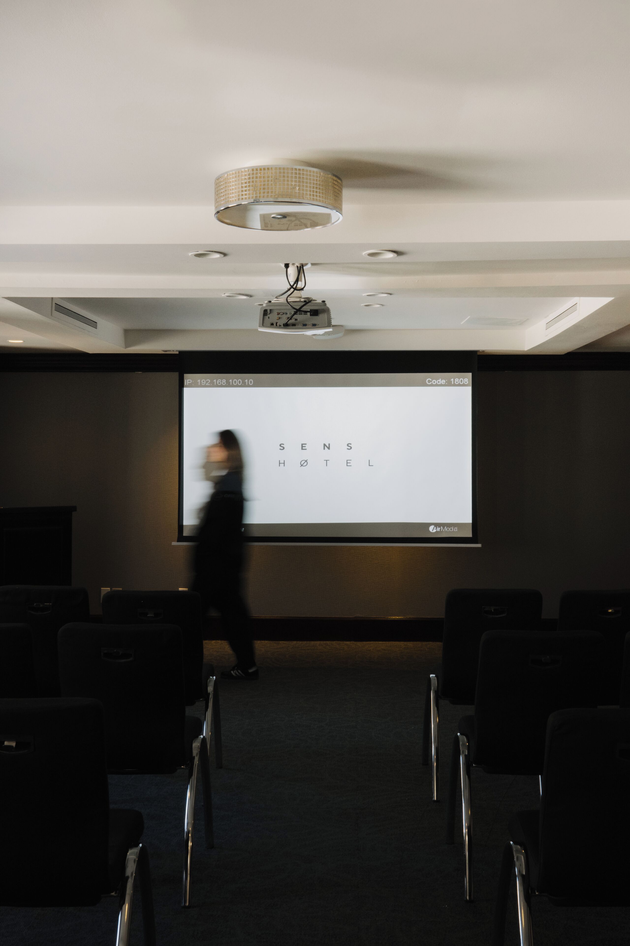 A projection screen with text, a ceiling light fixture, and rows of chairs in a room.