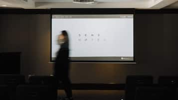 A projection screen with text, a ceiling light fixture, and rows of chairs in a room.
