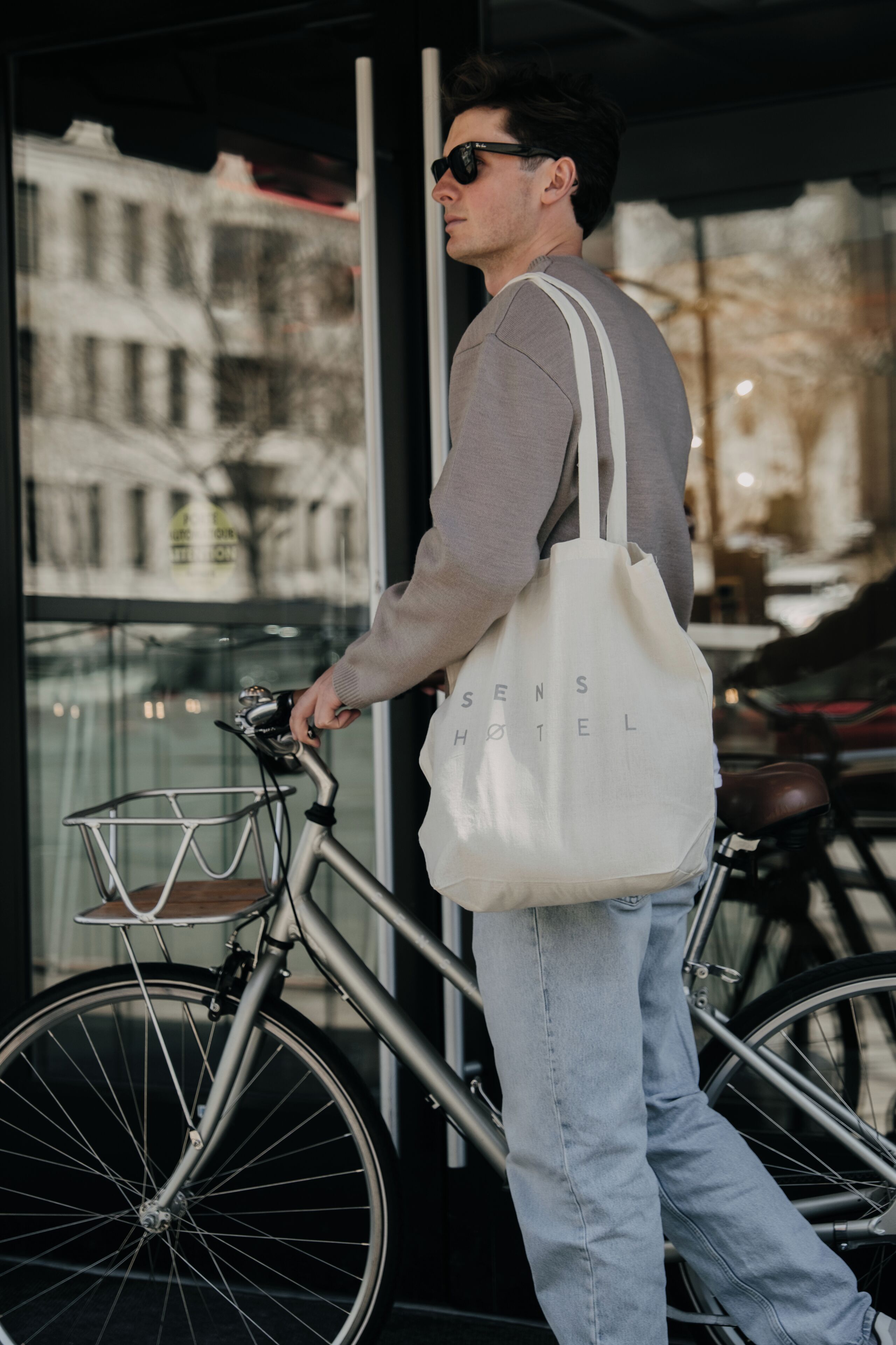 A person holding a bicycle and a white tote bag with 'SENS HOTEL' text, standing next to a bicycle with a basket.