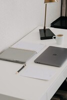 A white desk with a laptop, a notebook, a pen, a lamp, and a cup of coffee.