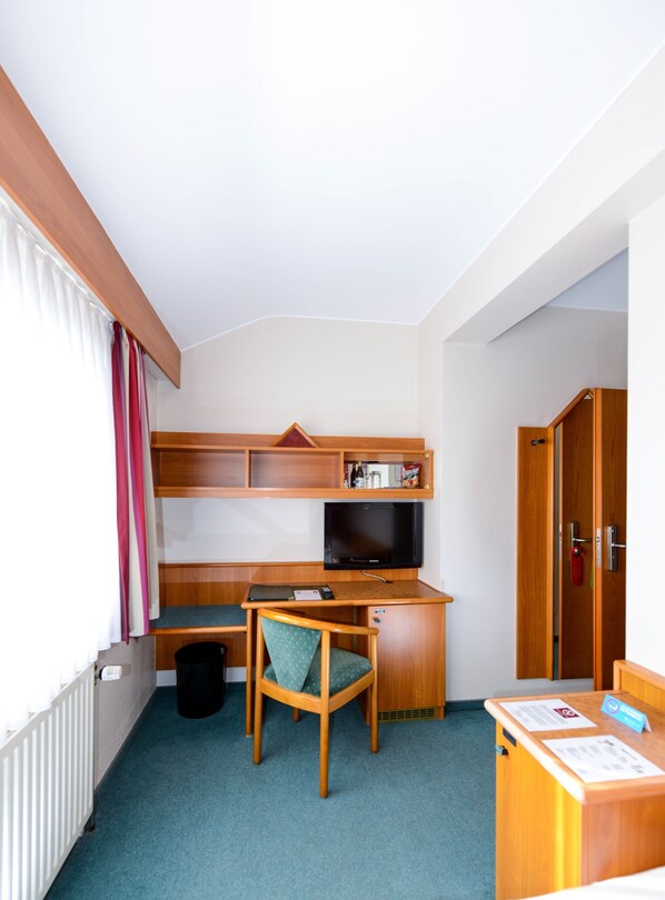 A compact hotel room with a wooden desk, a television, a chair, and a wardrobe.
