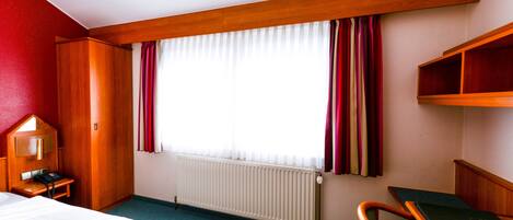 Een hotelkamer met een bed, een bureau, een stoel, een radiator en een raam met gordijnen.