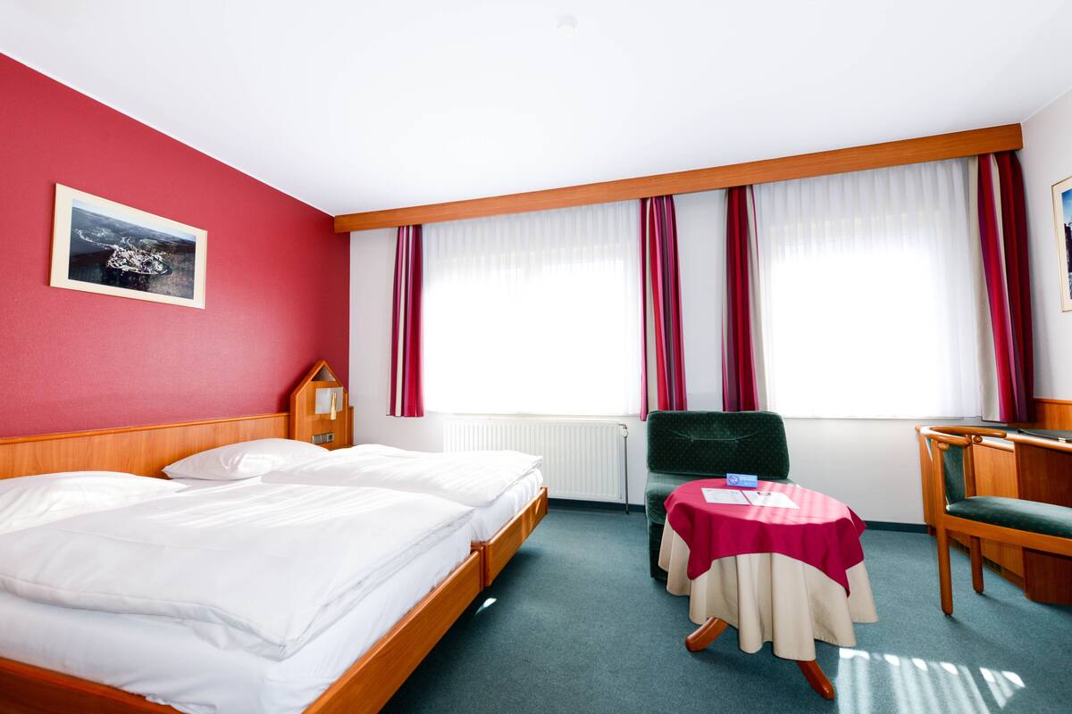 A hotel room with a bed, a desk, a chair, a table with a tablecloth, a radiator, and a window with curtains.
