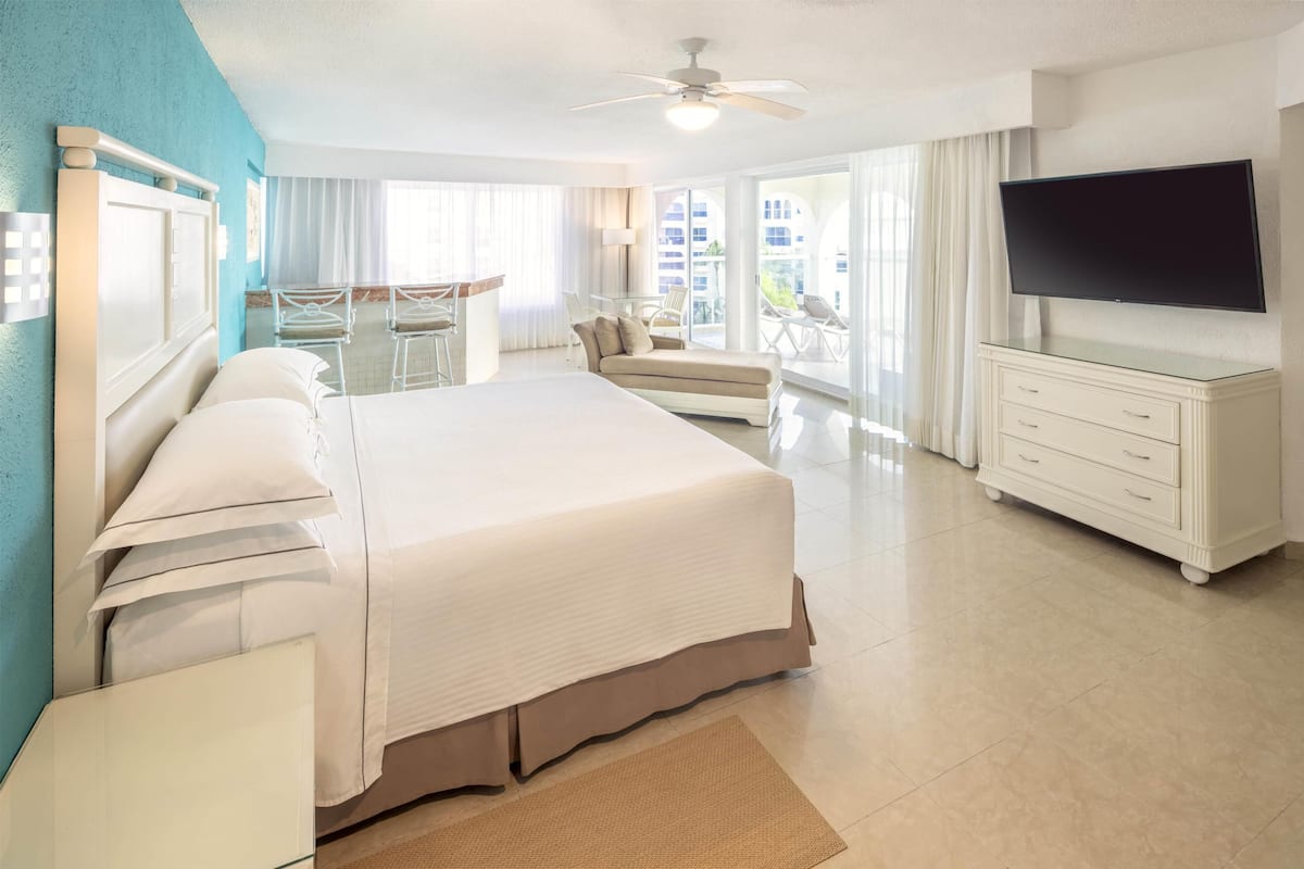 Habitación de hotel con una cama grande, televisión, escritorio y balcón con sillas.