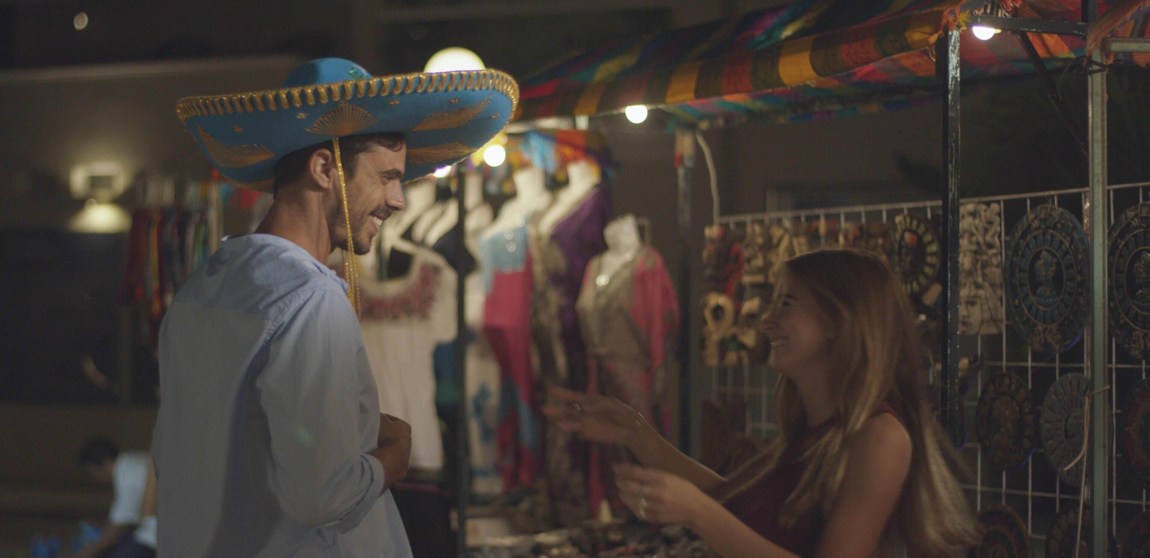 Ein Mann mit blauem Hemd und Sombrero steht an einem Marktstand mit verschiedenen Waren.