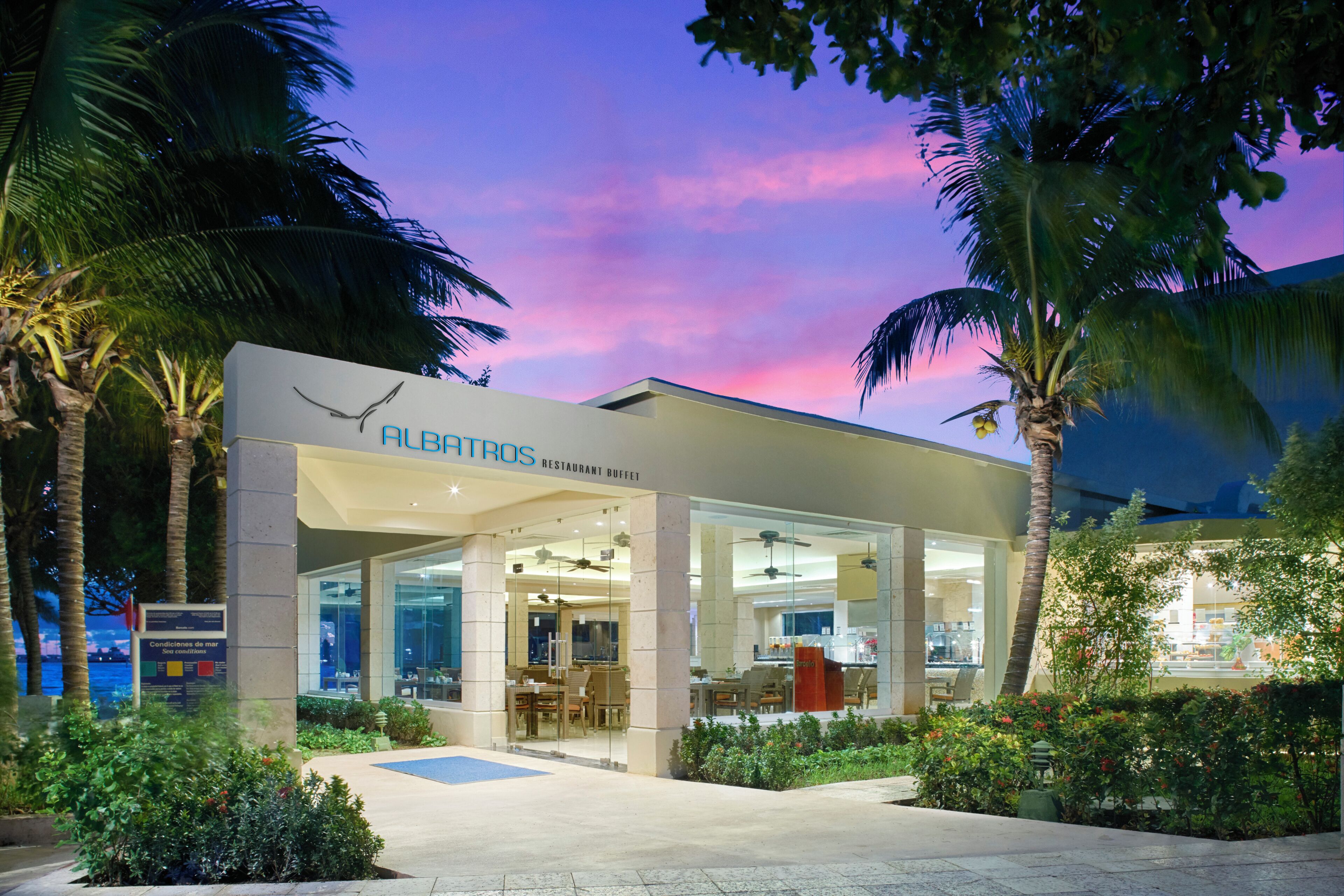 A restaurant with a glass facade, palm trees, and a view of the sky.