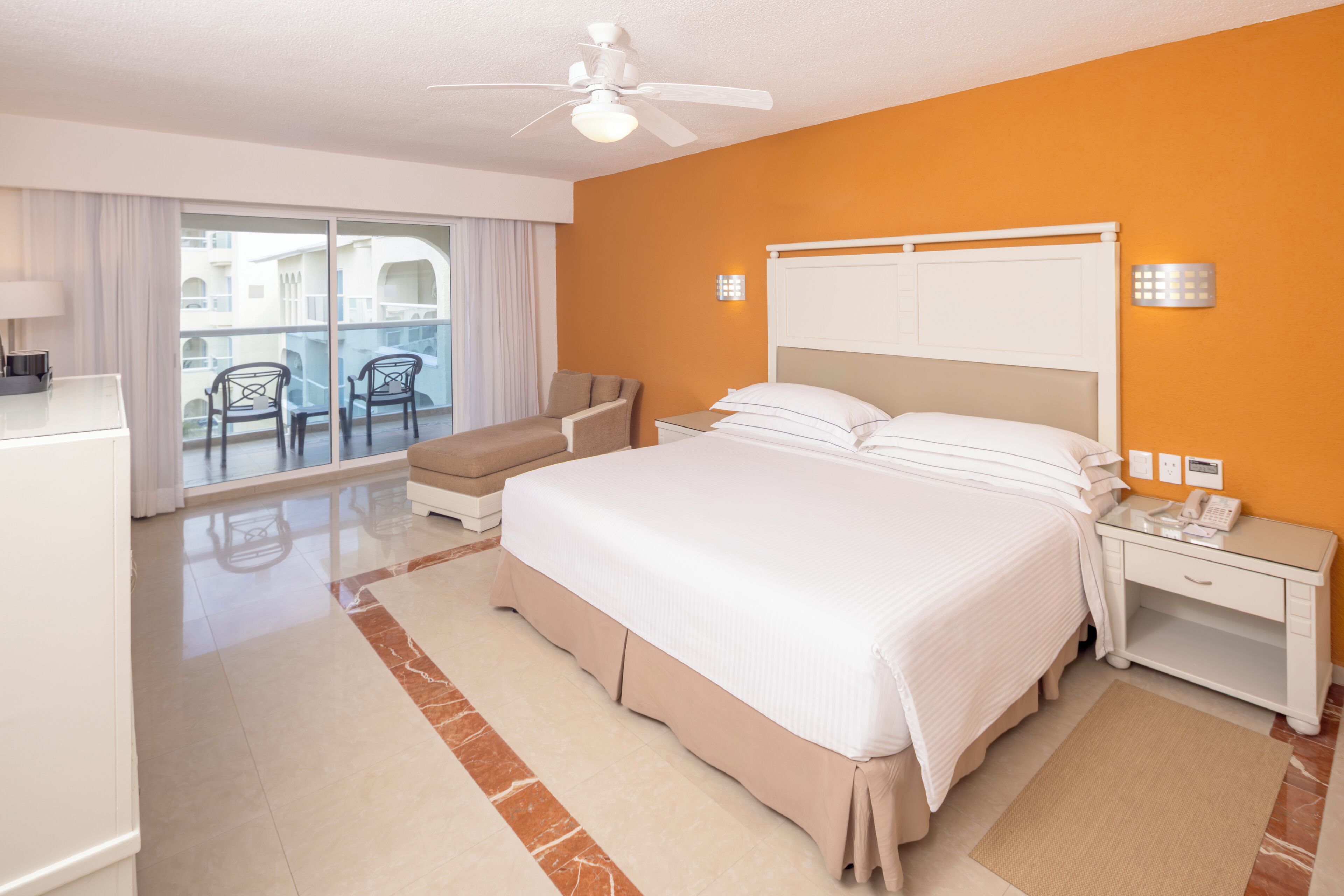 A hotel room with a bed, a desk, a chair, a balcony with chairs, and a ceiling fan.