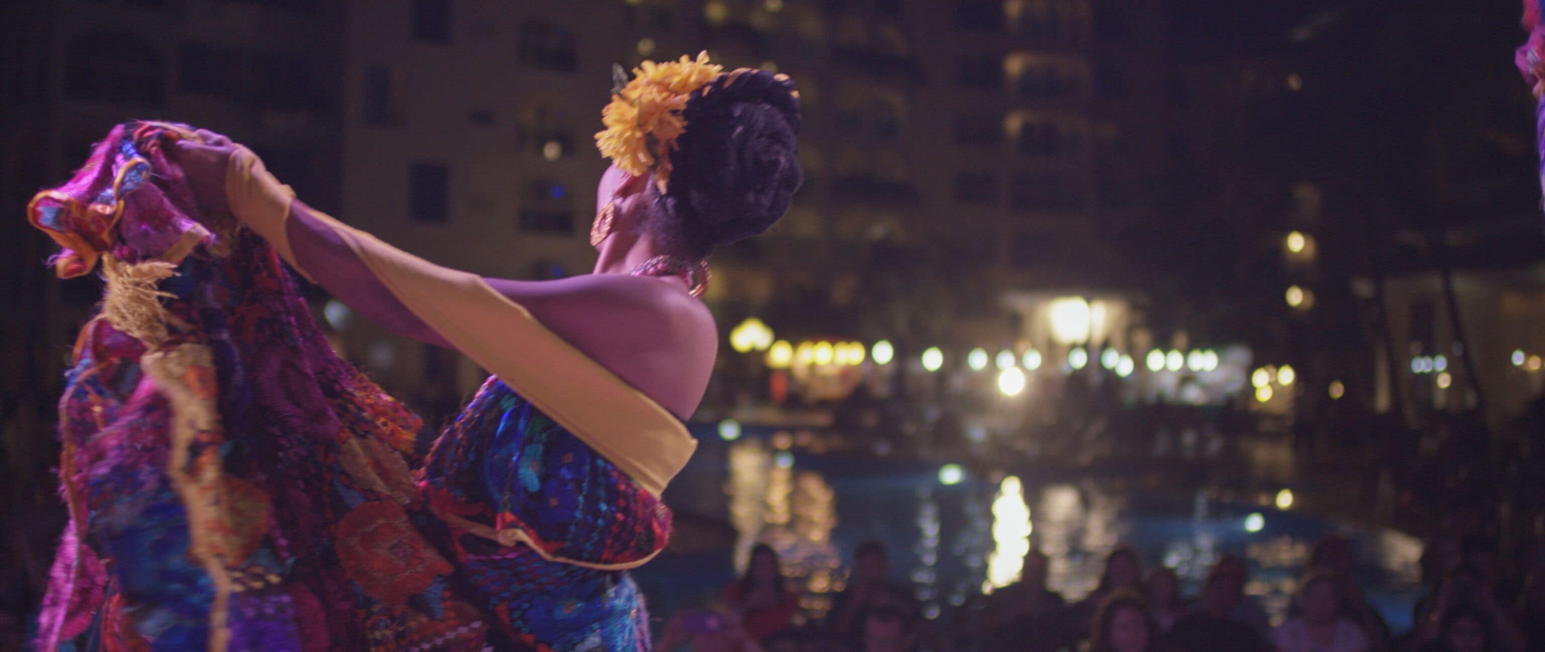 A person in a colorful costume with a headpiece, standing in a public space with a body of water and buildings in the background.