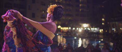 A person in a colorful costume with a headpiece, standing in a public space with a body of water and buildings in the background.
