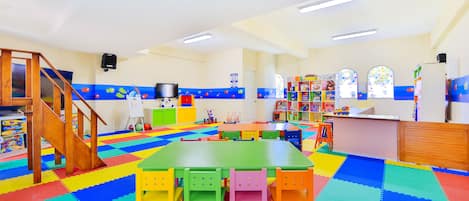 A brightly lit playroom with colorful mats, a green table, and chairs.