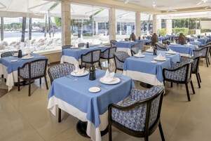Un restaurante con varias mesas y sillas, cada conjunto con platos, cubiertos y servilletas. Las mesas tienen manteles azules, y las sillas presentan un diseño geométrico. El restaurante tiene grandes ventanales que ofrecen vistas a una piscina y palmeras.
