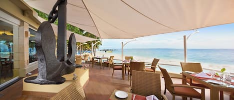 Un área de comedor al aire libre con mobiliario de mimbre, una gran escultura y vista a la playa y al océano.