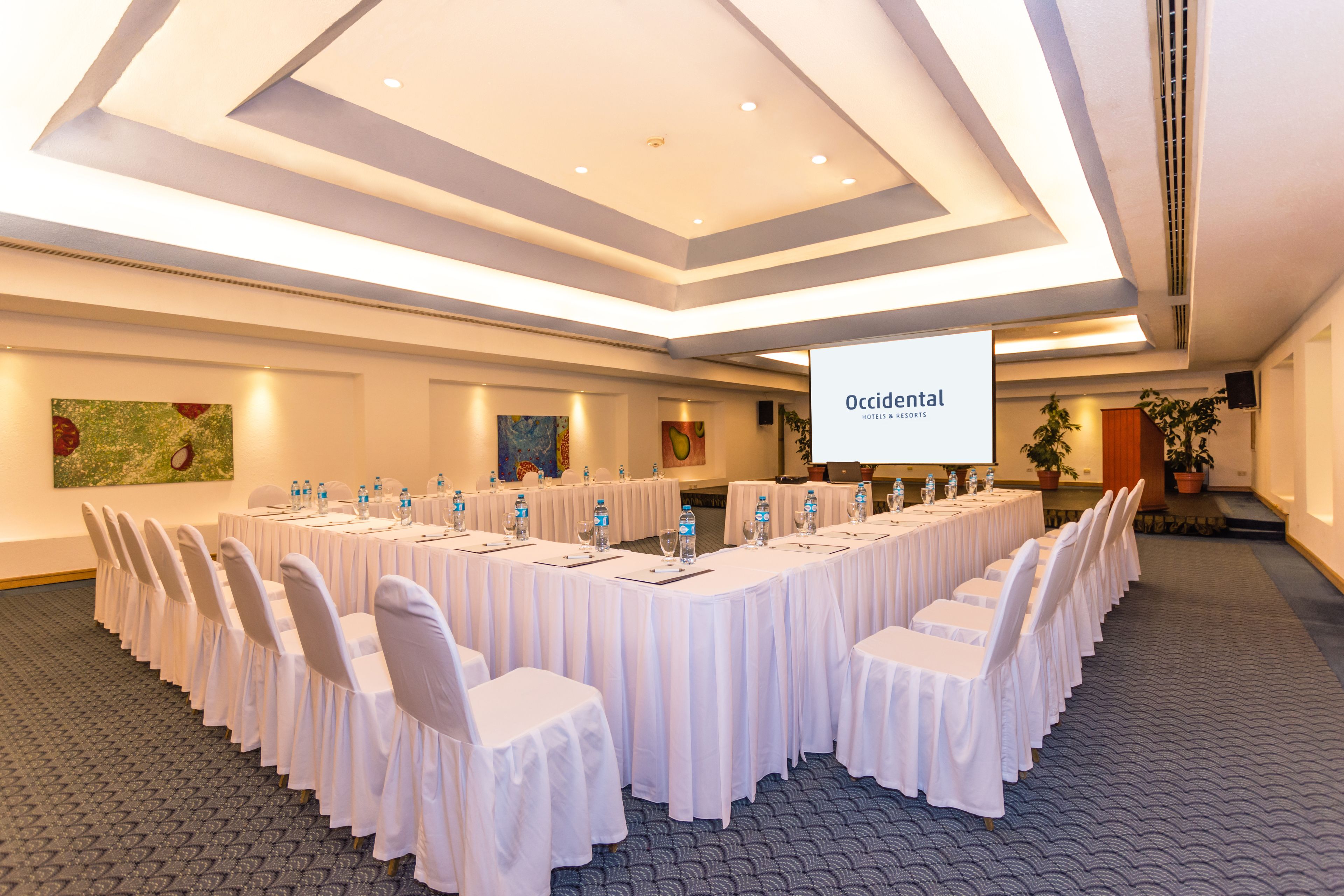Un salón de conferencias con una larga mesa lista para una reunión, sillas blancas y una pantalla de proyector mostrando 