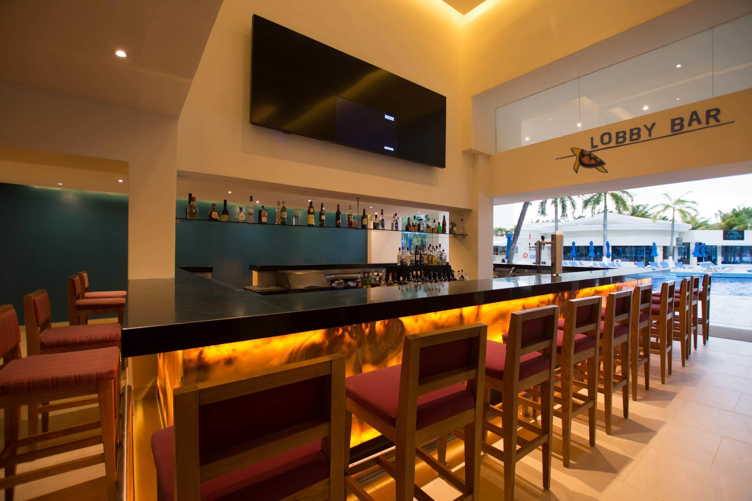 Zona de bar con una televisión de pantalla plana sobre la barra, mostrador con una luz cálida trasera, y un cartel que dice 