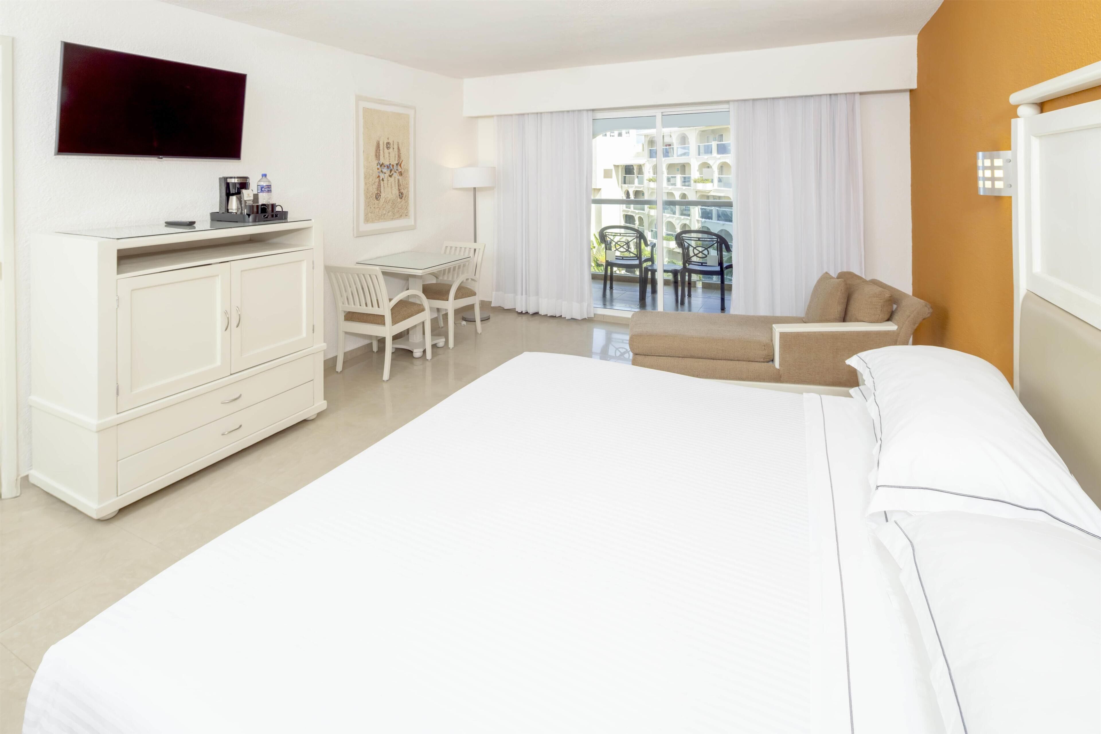 Une chambre d’hôtel avec un grand lit, un bureau avec des chaises, une télévision et un balcon donnant sur des immeubles.